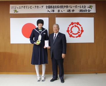 「JOCジュニアオリンピックカップ全国都道府県対抗中学バレーボール大会入澤まい選手激励会」と書かれた横幕が貼られ、国旗と三郷市市旗が飾られた室内で、右手にマスコットキャラクターかいちゃんの小さなぬいぐるみを持ち左手で市長と一緒に目録を持って立っている入澤さんの写真