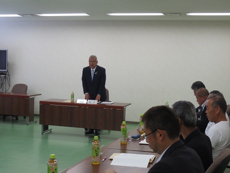 三郷市園芸協会総会で立って挨拶の言葉を述べる三郷市長と、席札とお茶のペットボトルが置かれた席に座り話を聞く参加者の写真