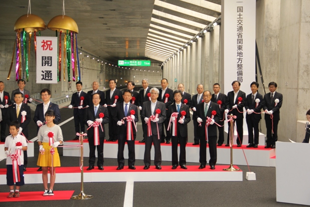 道路上に設置された特設ステージに立って胸章をつけた関係者や子どもたちがテープカットを行っている写真