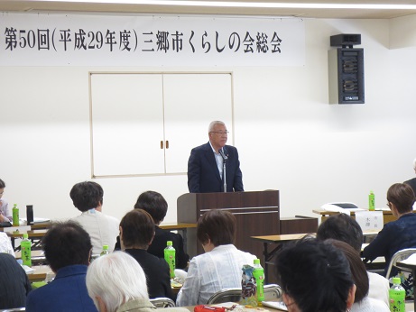 「第50回(平成29年度)三郷市くらしの会総会」と書かれたポスターが壁に貼り付けられてあり、演台に立って挨拶の言葉を述べる三郷市長とペットボトルのお茶が置かれた席に座り話を聞く参加者の写真