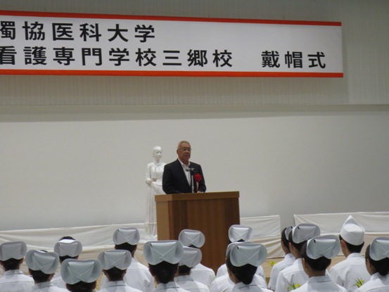 「獨協医科大学附属看護専門学校三郷校戴帽式」の吊り看板が設置された舞台上で、ナース服を身に着けた生徒たちの前で話をしている市長の写真
