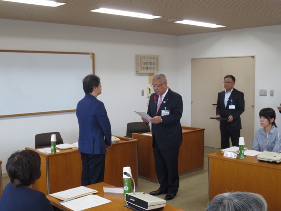 前に立ち子ども子育て対策の諮問書の内容を読み上げる三郷市長と、市長と向かい合って立ち話を聞く会長の男性の写真