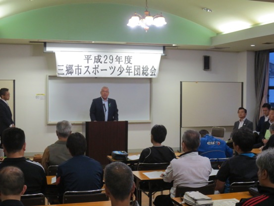 「 平成29年度三郷市スポーツ少年団総会」と書かれたポスターが天井に収納されているスクリーン下部に貼りつけられ、その下に置かれた演台に立ち話をする三郷市長と資料が置かれた席に座り話を聞く参加者の写真