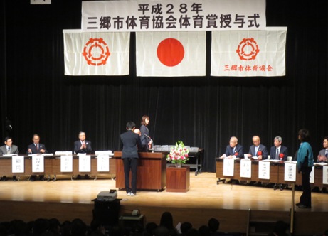 ステージ上の演壇の前で賞状を授与される男性と両側に配置された席に着席し授与の様子を見守っている関係者の写真