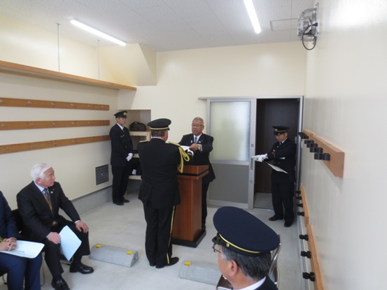 パイプ椅子が向かい合わせに設置された狭い機械器具置場に関係者たちが着席しているなか、入口近くに設置された演台を挟んで市長から本間雄一郎消防団長に目録を交付している様子の写真