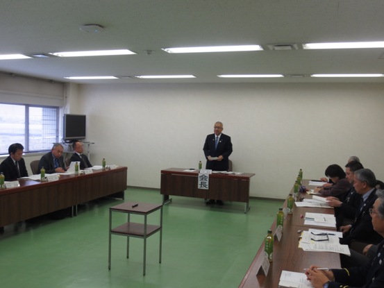 部屋の奥、中央に配置された、「会長」と書かれた席札が貼り付けられている席を立ち挨拶の言葉を述べる三郷市長と、部屋の両側に配置された席札とペットボトルのお茶が置かれている席に座り話を聞く関係者の写真
