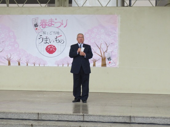 桜の木のイラストとともに「三郷春まつり桜とご当地うまいもの祭」と書かれた横断幕の前で、マイクを手に持ちステージ上で話をする三郷市長の写真