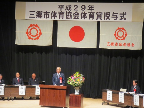 「平成29年三郷市体育協会体育賞授与式」と書かれた横断幕と国旗・三郷市旗・三郷市体育協会の旗が吊り下げられているステージ上で、赤・ピンク・黄色・白の豪華な花々で彩られた花台と並ぶ演台に立ち挨拶の言葉を述べる三郷市長と、ステージ上に配置された席札が付いている席に座り話を聞く来賓者の写真
