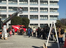 建物の前に消防車と、はしごが上に高く伸びたはしご車がとまっており、その前でたくさんの親子連れが1列に並んで順番待ちをしている様子の写真