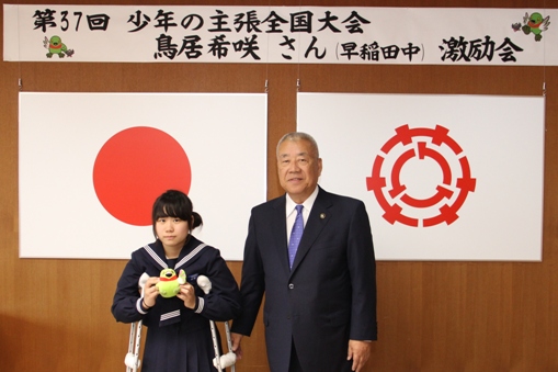 「第37回少年の主張全国大会 鳥居希咲さん(早稲田中)激励会」と書かれた横断幕と国旗と三郷市旗が飾られた室内で、マスコットキャラクターかいちゃんのぬいぐるみを持った鳥居さんと市長が並んで立っている写真