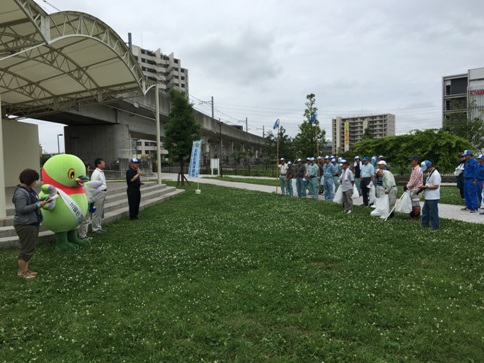 屋外ステージのある広場でごみ袋を持ちキャップを被っている方々と、ステージ前に手前から女性とマスコットキャラクターかいちゃん、男性と黒色のシャツを着た市長が向かい合って立ち開会式を行っている様子の写真