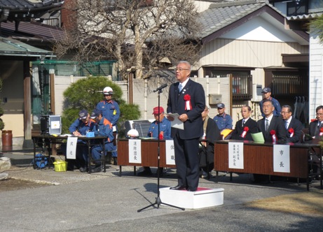 宝蓮寺の前に設置されたお立ち台に立ち挨拶の言葉を述べる三郷市長と、お立ち台の後ろに配置された席に座り話を聞く胸章をつけた住職と消防隊長、その他関係者の写真