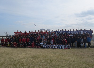 青地に「目指せゴール!がんばれ三郷FCJr.」と書かれている横断幕と赤い横断幕を持った参加者が最後列に横一列に並び、その前に選手たちが集まって並び、全員で記念撮影をしている写真