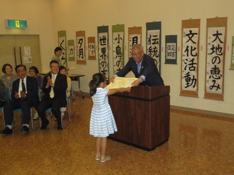 壁に書の作品が並んでいる室内で、奥に設置された椅子に座っている関係者が拍手を送るなか、演壇を挟んで向かい合って立つ市長が白色と水色のボーダー柄のワンピースを着た女の子に賞状を手渡している写真