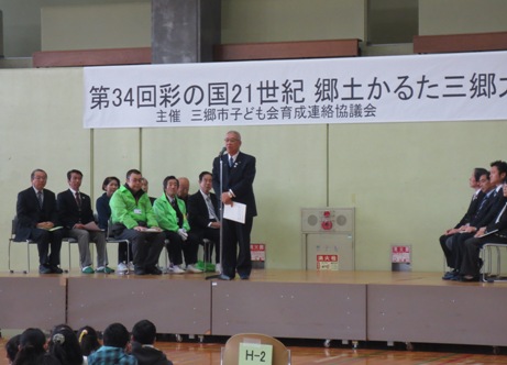 「第34回彩の国21世紀郷土かるた三郷大会」と書かれたポスターが貼りつけられている壁の前のステージに立ち挨拶の言葉を述べる三郷市長と、ステージ上に並べられたパイプ椅子に座り話を聞く関係者の写真