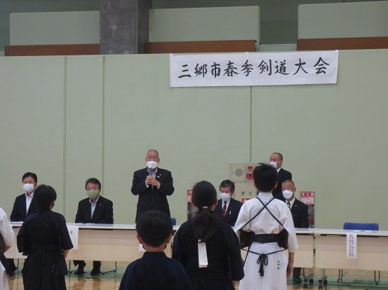 「三郷市春季剣道大会」と書かれた横断幕の前で席を立って挨拶する三郷市長と、防具を付けた子どもたちが立って整列して聞いている写真