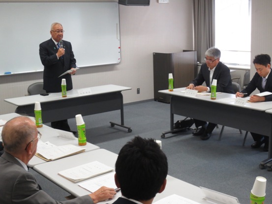 ホワイトボードの前に配置された席を立ちあいさつの言葉を述べる三郷市長と、席札とお茶のペットボトル・紙コップ・資料が置かれている席に座り話を聞く委員のメンバーの写真