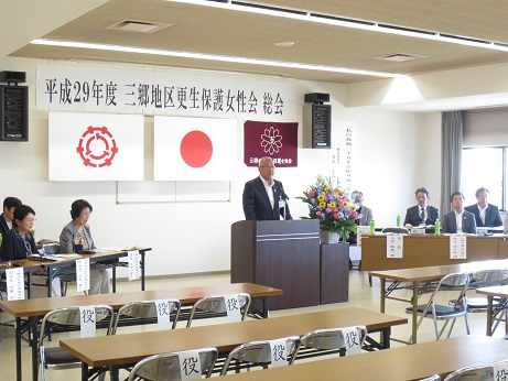 「平成29年度三郷地区更生保護女性会総会」と書かれたポスターの下には三郷市旗・国旗・更生保護女性会の旗が吊るされており、黄色・ピンク・水色の花々で彩られた花台と並ぶ演台に立ち挨拶の言葉を述べる三郷市長と演台の両側に設置された席に座り話を聞く関係者の写真
