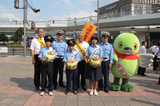 前列に3名の子どもが警察官の制服を着用しマスコットキャラクターかいちゃんのぬいぐるみを持ち、後列に市長と2名の警察官、黄色のタスキを肩にかけた男性とかいちゃんが並んでいる記念写真