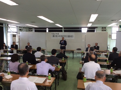 「平成27年度三郷市青少年育成市民会議総会」と書かれた横幕の席から立ちあがりマイクを片手に持っている市長の話を聞いている参加者の後ろ姿の写真