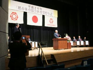 「平成26年三郷市体育協会体育賞授与式」と書かれた横断幕の下に国旗と三郷市市旗が飾られたステージの檀上で挨拶を行っている市長の写真