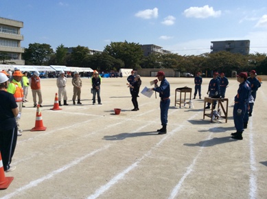 右手で拡声器を持ち資料を見ながら話しをしている男性の周りでヘルメットを被った参加者が聞いている写真