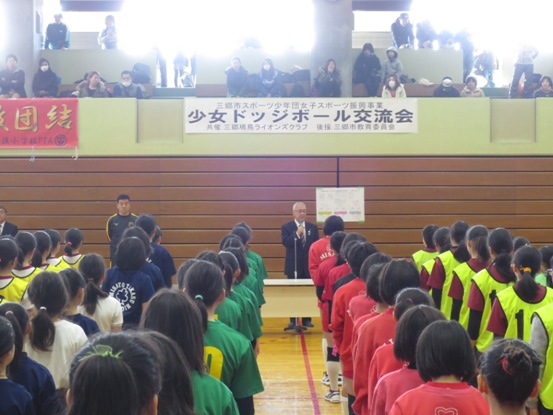 たくさんの観客がいる体育館の2階席から「少女ドッジボール交流会」と書かれた横断幕が掛けられている壁の前で、激励の言葉を述べる三郷市長と赤・緑・青などのユニフォームを着用した子どもたちがチームごとに整列し話を聞いている写真