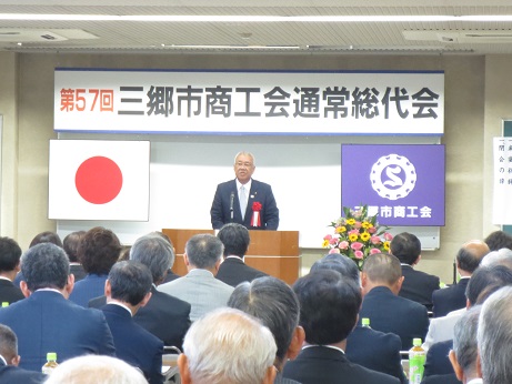「第57回三郷市商工会通常総代会」と書かれたボードと国旗、三郷市商工会の旗が吊り下げられ、ピンク・黄色の花々が飾られている花台の横にある演台に立ち話をする三郷市長と座って話を聞く大勢の参加者の写真