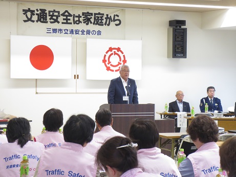 「交通安全は家庭から」と書かれた横断幕が壁に貼り付けられ国旗と三郷市旗が吊るされている会場で、演台に立って話をする三郷市長と話を聞く「Traffic Safety」のロゴがある薄ピンクのベストを着用した三郷市交通安全母の会メンバーの写真