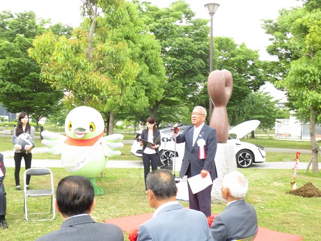 におどり公園の広場であいさつの言葉を述べる白い胸章を付けた三郷市長とパイプ椅子に座って話を聞く赤い胸章を付けた関係者、タスキをかけたかいちゃんと女性に抱かれているつぶちゃんの写真