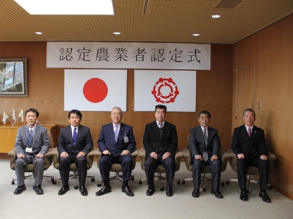 「認定農業者認定式」と書かれた横幕が壁に貼られた室内で、市長と認定式に参加した5名の関係者が横一列に並び座っている記念写真