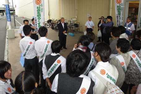 店舗に隣接する歩道上でスーツ姿の市長が「社会を明るくする運動啓発活動」と書かれたタスキを肩にかけた方々の前で話をしている様子の写真