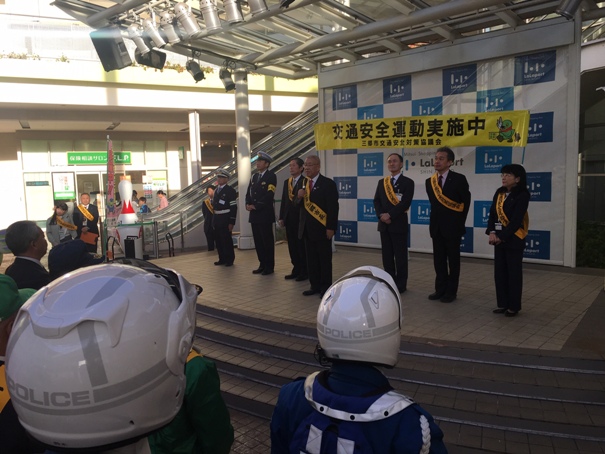 ステージに「交通安全運動実施中」の横断幕が掲げられ、ステージ上に6名の関係者が並んでいるなか、中央に立って話をしている市長と、ステージ下で市長の方を向いている関係者たちの写真