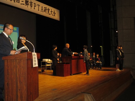 舞台上の手前側から、総合司会、表彰をする市長と受賞者、2列に並んだ椅子に座る来賓を写した表彰の様子
