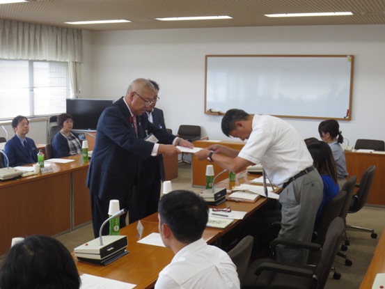 壁にホワイトボードが掛けられた室内で、委嘱書を手渡す三郷市長と深くお辞儀をしながら受け取る委員の男性、マイクと席札・お茶のペットボトルが置かれた席に座り交付の様子を見守る委員のメンバーの写真