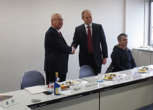 料理の乗った皿が並んだ白い長机の後ろに立った市長とルカス・カラツォリス特命全権大使が握手をして記念撮影をしている写真