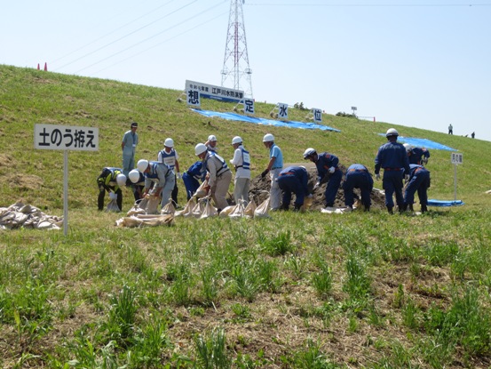 芝生の広がる土手に「土のう拵え」と書かれた看板が設置され、活動服とヘルメットを着用した関係者たちが土のうを拵えている写真