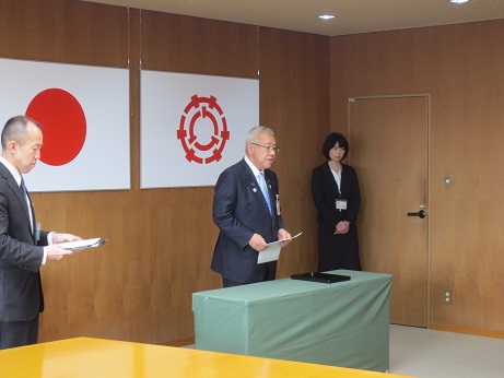 国旗と三郷市旗が吊り下げられている壁の前で、緑のカバーが掛けられ賞状盆が置かれた台の後ろに立ち挨拶の言葉を述べる三郷市長と、部屋の隅に立つ介添人の女性の写真