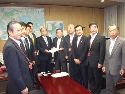 中央に立っている2名の男性が一緒に要望書を持ち、各市町の長と環境省の福山政務官、総務省の二之湯副大臣や関係者がテーブルを囲んで立っている写真