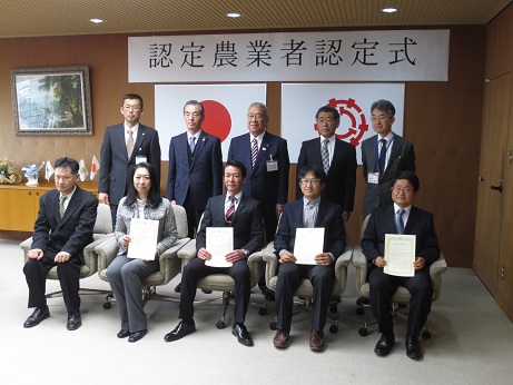 「認定農業者認定式」と書かれたパネルが張られた壁を背に、椅子に座り認定証を正面に向けた認定農業者の方々と、その後方で市長を中心に立つ関係者の集合写真
