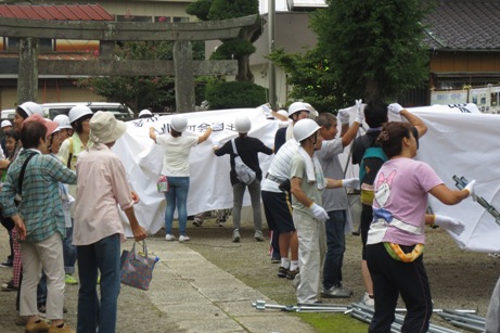 鳥居の手前の境内で、2組に分かれたヘルメットを被った参加者がテントを組み立てている訓練の様子の写真