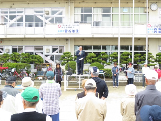 高州東小学校の校庭に設置された朝礼台に立ち話をする三郷市長と、向かい合って立ち話を聞く帽子を着用したゴルフ大会の参加者の写真