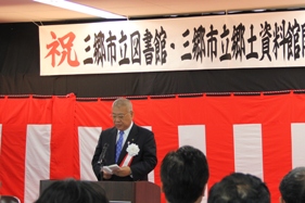 「三郷市立図書館・三郷市立郷土資料館開館式典」と書かれた横断幕の下の演壇に立ち挨拶を行っている市長の写真
