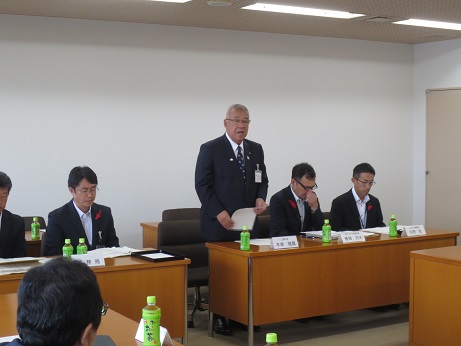 席を立ち挨拶の言葉を述べる三郷市長と、席札とお茶のペットボトルが置かれた席に座り話を聞く関係者の写真