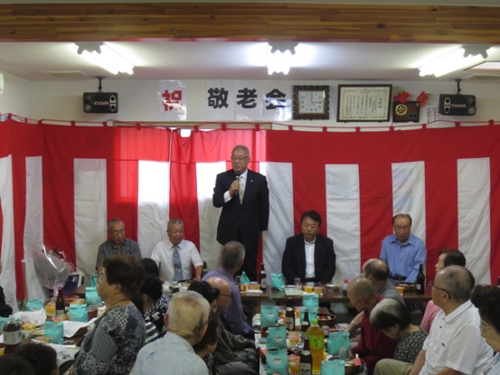 紅白の横断幕に囲われ、長机には飲み物や料理が用意された部屋で、多くの参加者たちが座って見守る中、市長が立って挨拶をしている敬老会の写真