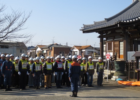 白いヘルメットと黄色と赤のゼッケンを着用した大勢の訓練参加者が宝蓮寺の前に整列している写真