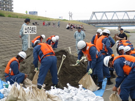 橋の手前の堤防でヘルメットに作業着を着用した作業員が「土のう拵え」と書かれたプラカードの傍に山積みになった土をスコップで土嚢袋に詰め、手前に口を縛って並べて置いている訓練の様子の写真