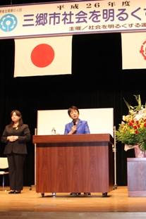 真っ直ぐ立っているスーツ姿の女性の右側に設置された檀上で右手でマイクを持ち講演を行っている幸島美智子先生の写真