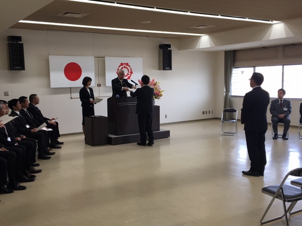日本国旗と三郷市章が掲げられた部屋で、関係者たちが両脇から見守る中、演台に立つ市長から向かいに立つ職員に辞令を渡している様子の写真