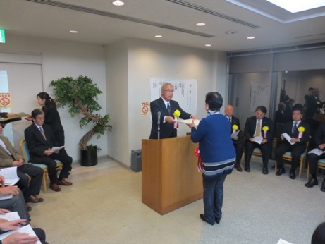 室内の壁側に設置された椅子に関係者が座り、中央の演壇を挟んで女性が市長から表彰状を手渡されている様子の写真